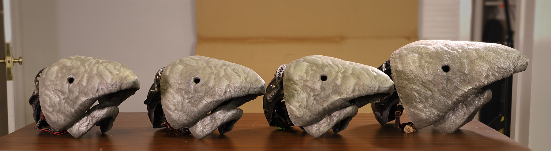 Four 3D-printed creature masks mounted on hardhat helmets, lined up on a workbench